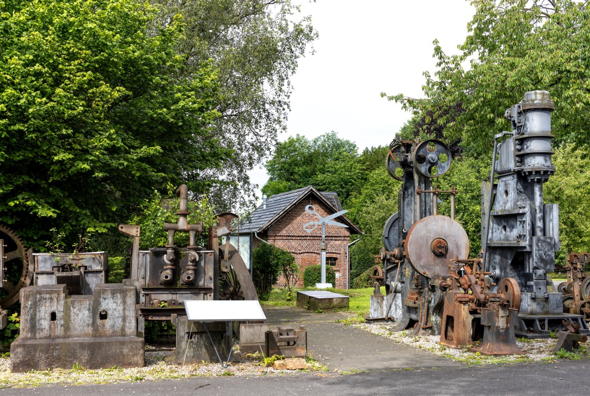 Alte Maschinen vor der Gesenkschmiede mit einer Informations-Tafel. Im Hintergrund ist die Statue einer Schere zu sehen. 