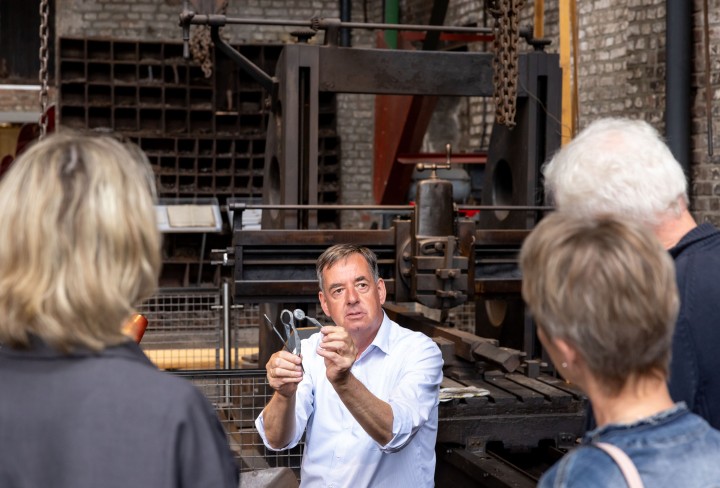 Männer und Frauen bekommen vom Museumsschmied seine Arbeit an einer Maschine erklärt