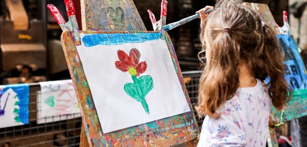 Kind steht mit dem Rücken zum Betrachter an einer Staffelei und malt mit einem Prinsel eine Blume. Im Hintergrund ist das Museum zu erkennen. 