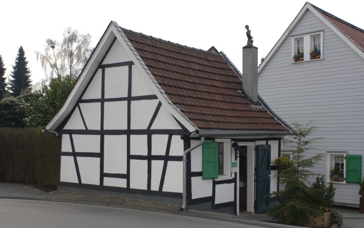 Fachwerkhaus mit weißer Fassade und dunklen Balken, grünem Fensterladen und rotem Ziegeldach an einer Straße.