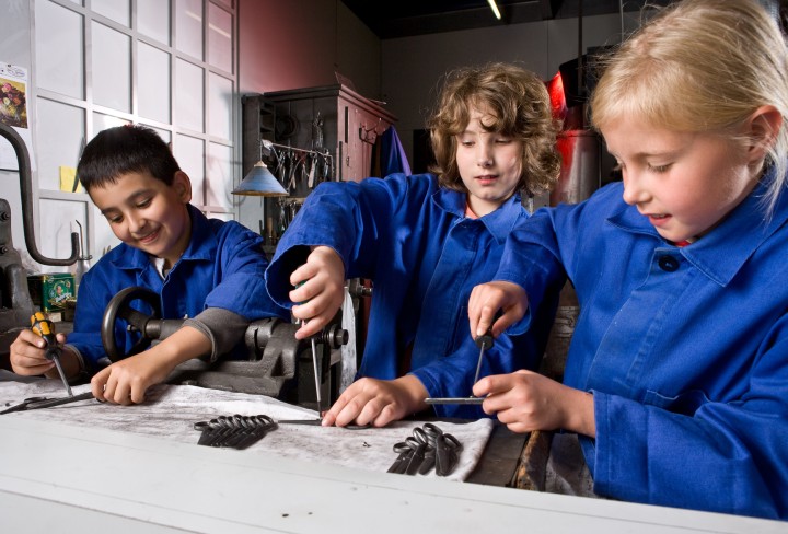 Ein Mädchen und zwei Jungen, in blaue Schutzkleidung gekleidet, stellen in der Scherenfabrik ihre eigene kleine Schere her.