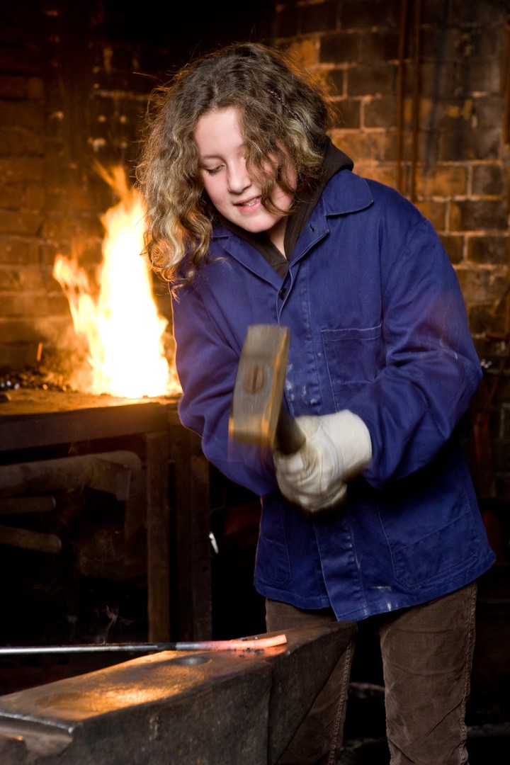 Ein Mädchen, in einer blauen Schutzjacke, haut mit einem Hammer auf ein glühendes Stück Eisen. Im Hintergrund lodert ein Feuer.