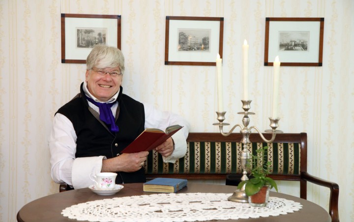 Hüttendirektor Gottlob Jacobi in weißem Hemd, schwarzer Weste und blauem Halstuch sitzt an einem runden Tisch mit weißer Spitzen-Tischdecke, hält ein aufgeschlagenes Buch und sitzt vor einer gepolsterten Holzbank mit gestreiftem Bezug.
