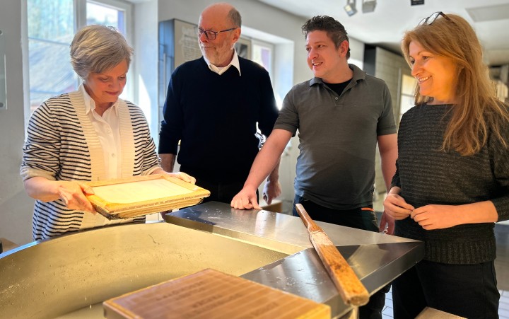 Vier Erwachsene stehen gemeinsam um eine Schöpfbütte, eine Frau hebt einen Schöpfrahmen aus der Flüssigkeit heraus