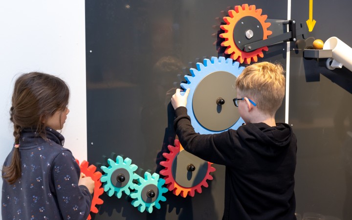 Zwei Kinder bedienen bunte Zahnräder an einer Wandinstallation, ein Junge dreht ein großes blaues Zahnrad.