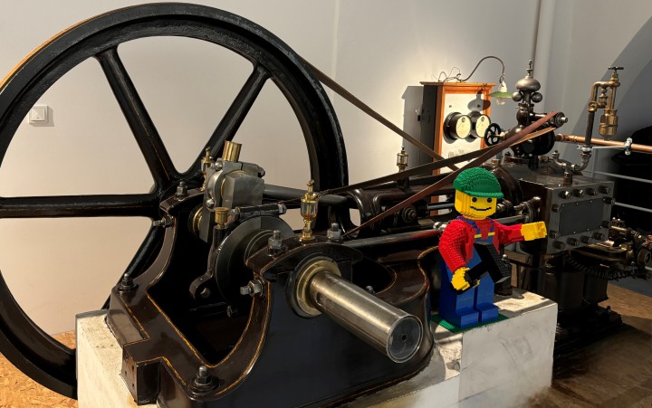 Große historische Maschine mit Schwungrad und einem Lego-Männchen in rotem Oberteil und grüner Mütze auf einem Sockel daneben.