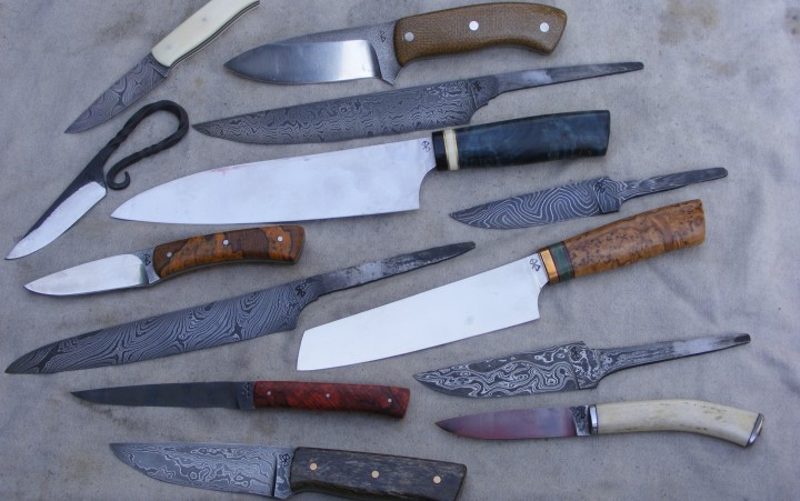 Mehrere handgefertigte Messer mit unterschiedlichen Klingenformen und Griffen aus Holz und anderen Materialien auf grauem Untergrund angeordnet.