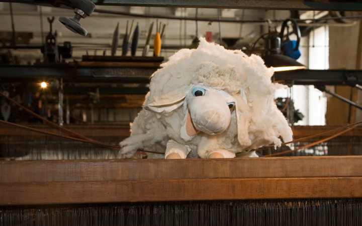 Ein Plüschschaf mit weißem, flauschigem Fell und blauen Augen ist auf einem Holzbalken vor einer Webmaschine zu sehen.