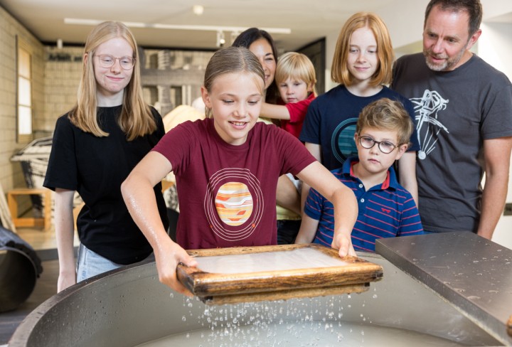 Eine Personengruppe, bestehend aus Kindern und Erwachsenen, bei dem ein Mädchen auf altmodische Art Papier aus Wasser schöpft.