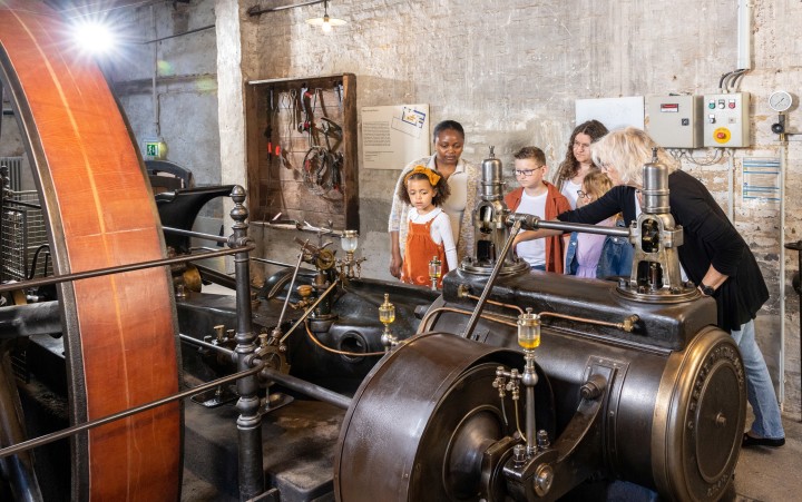 Gruppe von Kindern und zwei Erwachsene steht neben einer großen historischen Dampfmaschine in einer Werkstatt