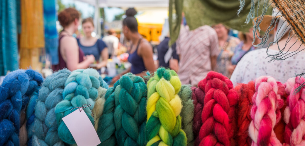 Bunte Wolle hängt geflochten und nach Farben sortiert im Vordergrund. Im Hintergrund ist Markttreiben zu sehen. 