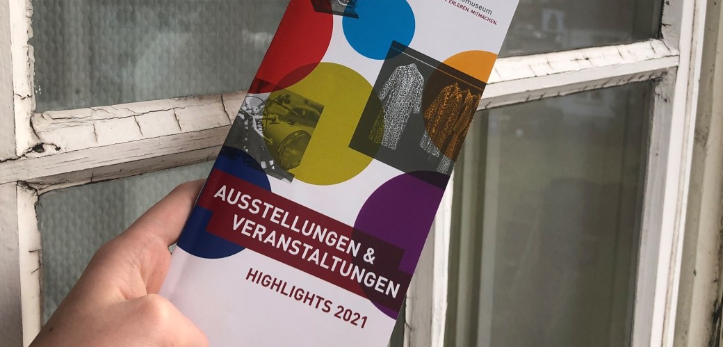 Eine Hand hält einen bunten Kalender mit der Aufschrift: Ausstellungen & Veranstaltungen, Highlights 2021, im Hintergrund spiegeln sich Bäume in einer Fensterscheibe
