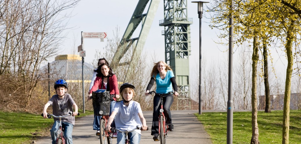 Zwei Frauen und zwei Jungen fahren auf Fahrrädern einen kleinen Hügel hinunter. im Hintergrund sieht man einen Förderturm von einer Zeche.