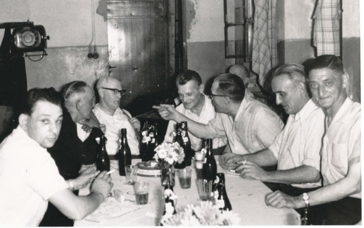 Schwarz-weiß-Fotografie von mehreren Männern, die an einem Tisch mit Flaschen und Gläsern sitzen und sich unterhalten.