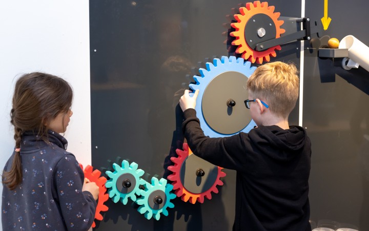Zwei Kinder bedienen bunte, große Zahnräder an einer Wandinstallation, eine Kugel rollt auf einer Schiene.