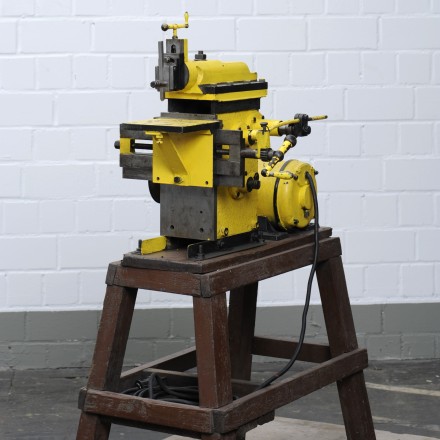 Eine kleine, gelb lackierte Shapingmaschine. Aufgestellt auf einem Holzbock.