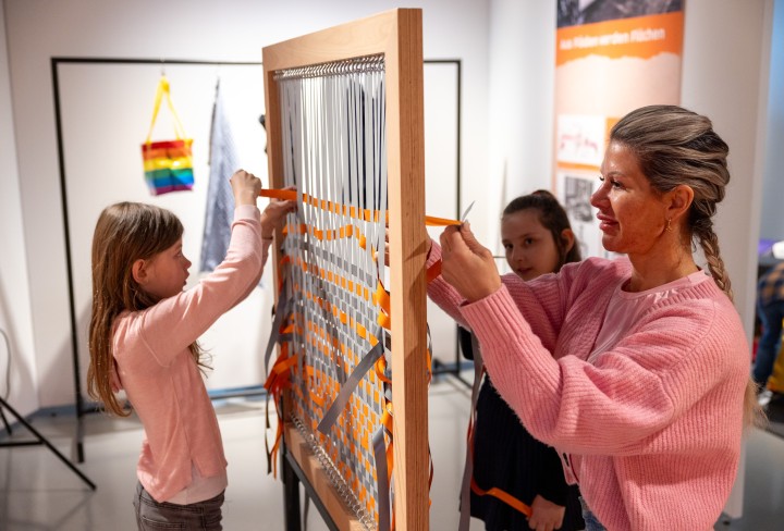 Eine Frau und zwei Mädchen tüfteln an einem Webrahmen in einer Ausstellung